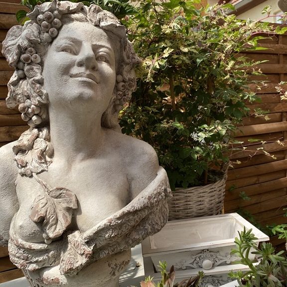 Skulptur einer lächelnden Frau mit Blumen im Haar, umgeben von Pflanzen und einem Topf.