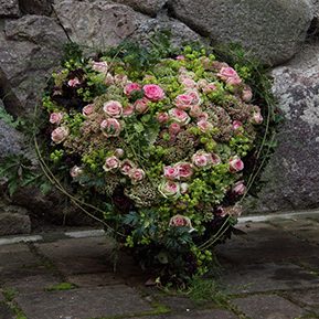 Herzförmiger Blumenarrangement mit rosa und grünen Blüten vor einer steinernen Wand.