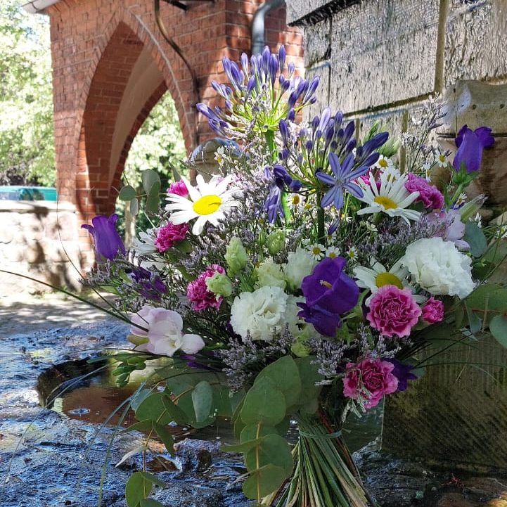Bunte Blumenarrangement mit weißen, lila und rosa Blüten vor einer historischen Steinmauer.