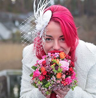 Frau mit pinken Haaren hält einen bunten Blumenstrauß und trägt einen eleganten Kopfputz.