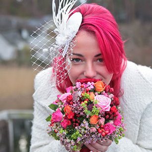 Frau mit pinken Haaren in Hochzeitskleidung hält einen bunten Blumenstrauß vor sich.
